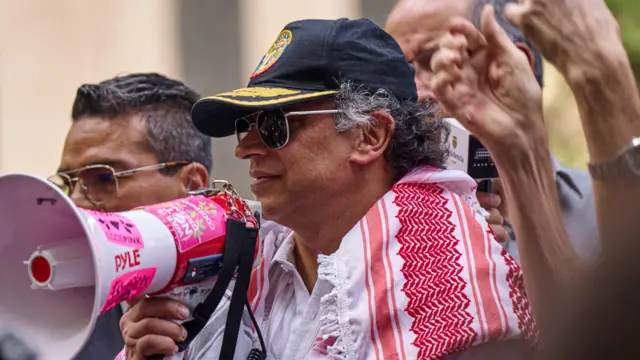 Gustavo Petro com um megafone na mão em uma assembleia pró-palestina em Nova York. Ele está de bone azul, óculos escuros, camisa branca e um tecido bordado vermelho e branco nas costas