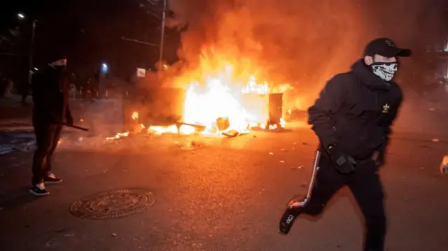 Zamaskowany protestujący w czapce baseballowej ucieka przed dwoma płonącymi kontenerami na śmieci. W tle widać szereg policjantów uzbrojonych w tarcze.