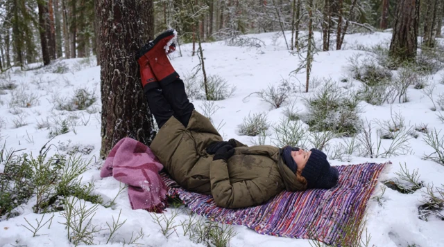Kobieta w zimowym płaszczu i czerwonych kaloszach leży na wzorzystym kocu rozłożonym na ściółce leśnej. Nogi opiera o drzewo. Wokół niej widać ośnieżony zimowy las. 