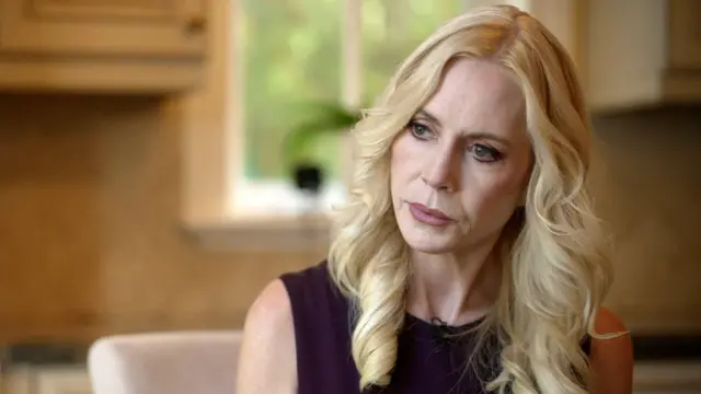 ウェーブのかかったブロンドの髪に黒いドレスを着たマコーリー氏が、オフィスの室内でクローズアップで写っている。カメラの左方向を見つめ、背景はぼやけている