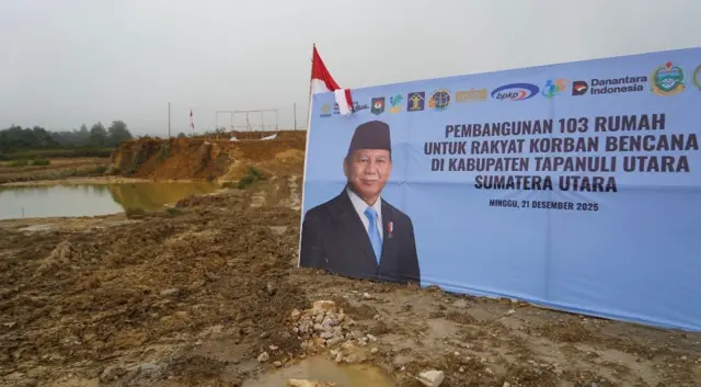 Lahan kosong dengan genangan air terlihat di kejauhan terlihat di sebelah kiri, sementara di sebelah kanan ada spanduk besar bergambar foto Presiden Prabowo dengan tulisan, "Pembangunan 103 rumah untuk rakyat korban bencana di Kabupaten Tapanuli Utara, Sumatera Utara. Minggu, 21 Desember 2025". 