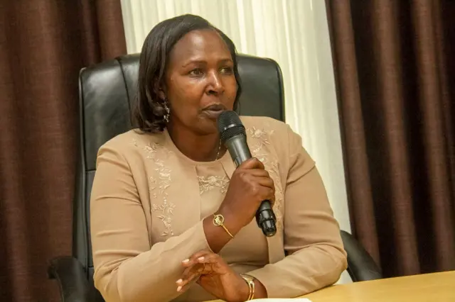 Umugore wambaye imyenda ya kakhi, afite indangururamajwi mu ntoki asa n'urimo kuvuga ijambo.