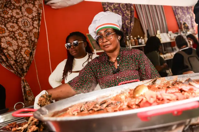Women stand behind a large pot of food.