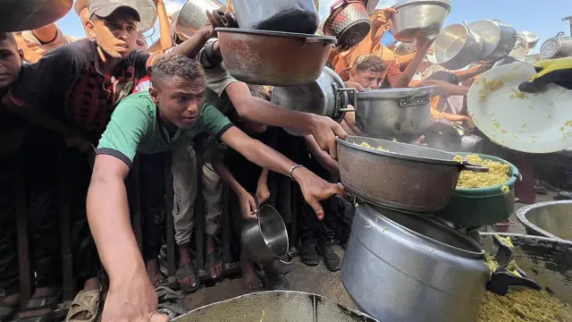 A crowd of Palestinians reach out their hands with empty pots to receive hot meals distributed by charity organisations in the al-Mawasi area.