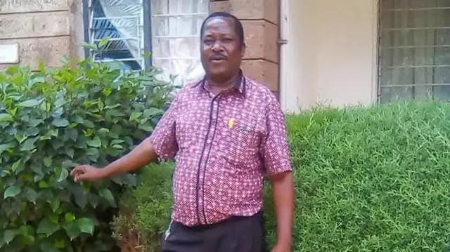 Benjamin Mutuku na tsaye a gabanin gidansa. Ya na sanye da riga taguwa mai gajeren hannu, ya dafa wata fulawa da ke kusa da inda yake tsaye.