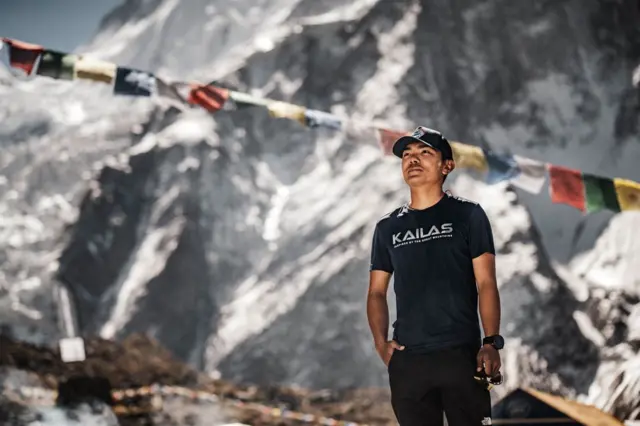 A man dressed in black, wearing a black cap, stands against the backdrop of a snow-covered, icy mountainside