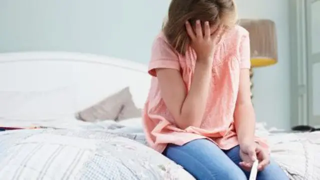 Молода дівчина у джинсах і рожевій блузці сидить на ліжку. В одній руці тримає тест на вагітність, іншою прикриває обличчя.