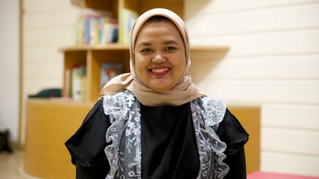 Mimma mengenakan jilbab warna cokelat, baju hitam dengan rompi renda berwarna putih. Dia tersenyum ke arah kamera.