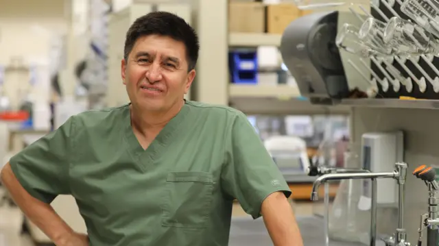 Homem vestindo camisa verde-oliva com equipamento de laboratório ao fundo, sorrindo para a câmera