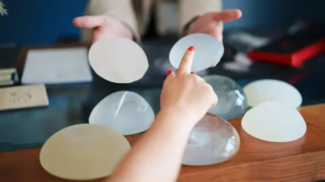 Kobieta z czerwonymi paznokciami wskazuje na protezę piersi, którą podaje jej inna osoba trzymająca dwie protezy. Na drewnianym stole leży sześć protez piersi
