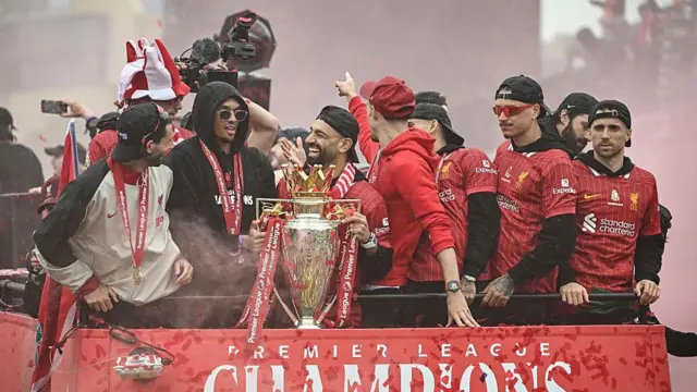 بازیکنان تیم لیورپول با لباس‌های سرخ که جام قهرمانی را در دست دارند و خوشحال هستند.