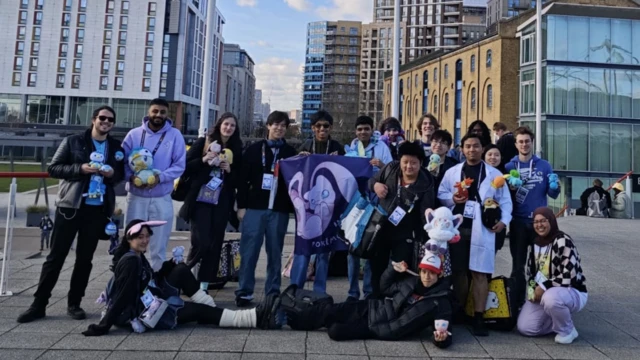 صورة تضم 17 عضواً من جمعية بوكيمون بجامعة لندن، يقفون في الهواء الطلق أمام مبانٍ شاهقة في المدينة. يحمل كل منهم دمية بوكيمون أو قطعة من البضائع التي تشير إلى شخصيتهم المفضلة.