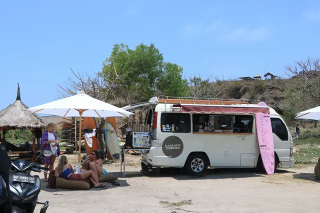 Mobil van berwarna putih yang difungsikan sebagai warung kopi kecil diparkir di pantai. Di sebelahnya ada tiga orang turis asing yang sedang bersantai di bawah payung pantai yang cukup besar.
