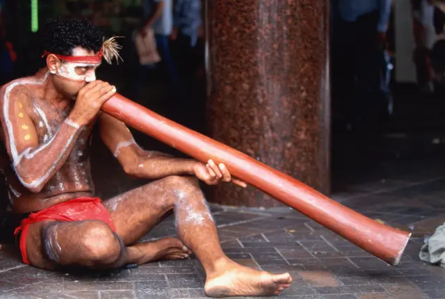 Un aborigen australiano, en taparrabos rojo, con la cara y cuerpo pintados de líneas y puntos blancos, está sentado soplando por un tubo largo de caña