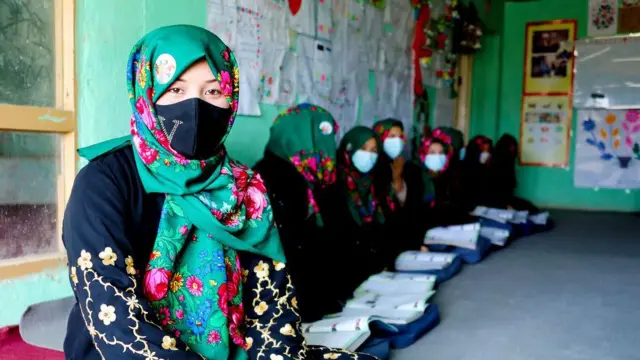 تصویر شماری از زنان بازگشت‌کننده در مکانی شبیه صنف درسی. در عکس زنی جوانی دیده می‌شود که چادر سبز گلدار به سر دارد ولباس سیاه پوشیده است. این زن به سوی دوربین می‌بیند اما صورتش با ماسک پوشانده شده‌است. 