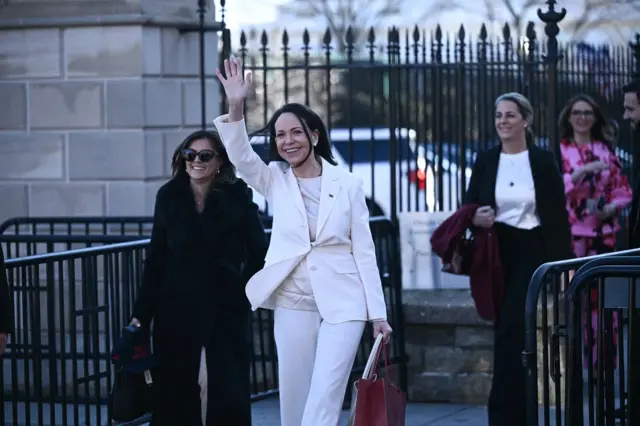 María Corina Machado, vestida de blanco y con una cartera marrón, saluda. A su lado hay una mujer con lentes de sol, vestida de negro y atrás, otra mujer con una blusa blanca y un traje negro.