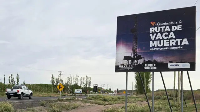Estrada por onde passa uma caminhonete, com um cartaz dando as boas-vindas a Vaca Muerta