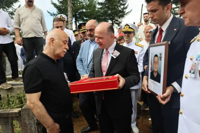 Bir kişi şehitlik belgesinin olduğu kutuya bakıyor. Bir görevli hayatını kaybeden askerin fotoğrafını taşıyor.