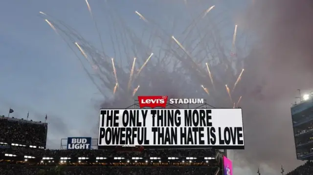 夕暮れの空を背景に花火が打ち上げられ、掲示板では白地に黒いブロック体の大文字で「THE ONLY THING MORE POWERFUL THAN HATE IS LOVE」と書かれている