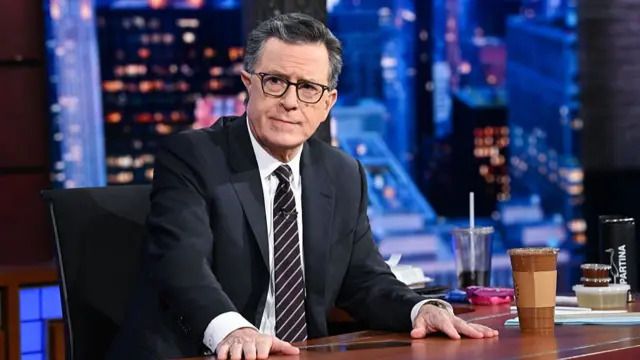 Stephen Colbert sentado atrás de uma mesa em um estúdio de televisão, usando terno escuro, camisa branca, gravata listrada e óculos. Ele apoia as mãos sobre a mesa e olha levemente para o lado com expressão séria. Ao fundo, aparecem luzes desfocadas de uma paisagem urbana noturna, enquanto sobre a mesa há copos de bebida, papéis e outros objetos de trabalho.