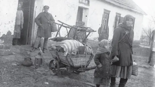 Старе чорнобіле фото, на якому зображена родина біля будинку, на першому плані - літня жінка з дитиною, за нею віз з речами