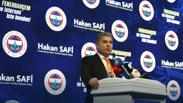 Mikrofonda konuşan bir adam. Arkada Fenerbahçe logosu ve Hakan Safi ismi görülüyor.