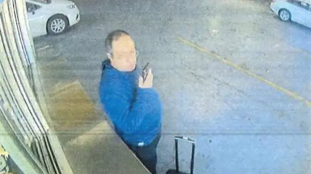 Uma pessoa segurando um telefone celular e vestindo um moletom azul aparece ao lado de uma mala com rodinhas, em um estacionamento subterrâneo, com carros ao fundo.
