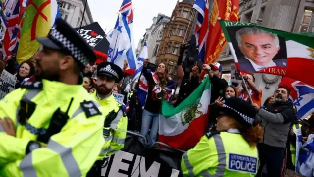 Женщина, укутанная флагом Великобритании, стоит в толпе за линией полицейских в ярких жилетах. Она указывает на людей, стоящих за камерой.