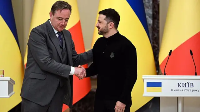 بارت دی ویور، نخست‌وزیر بلژیک و ولودیمیر زلنسکی، رئیس‌جمهور اوکراین در حال دست دادن مقابل پرچم‌های دو کشور بلژیک و اوکراین