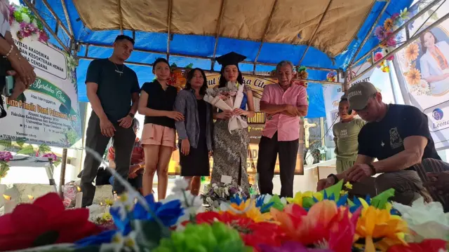 Tiara Maharani Dwi Pakaenoni adik dari dokter Icha berdiri di sisi pusara kakaknya saat mengenakan toga dalam perayaan wisuda sarjana kedokteran. 