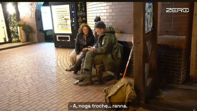 Двое людей сидят на скамейке на улице с брусчаткой. Мужчина одет в камуфляжную форму и держит в руках телефон; рядом на земле стоит большой рюкзак и прислоненная трость. Женщина сидит рядом и разговаривает с ним. Сзади видны кирпичные стены, освещенная витрина и елочные гирлянды. В нижней части изображения показан субтитр: «А, нога немного… ранена».