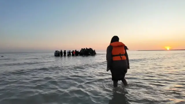 یک پناهجو به پناهجویانی که سعی می‌کنند سوار قایق بادی شوند نگاه می‌کند