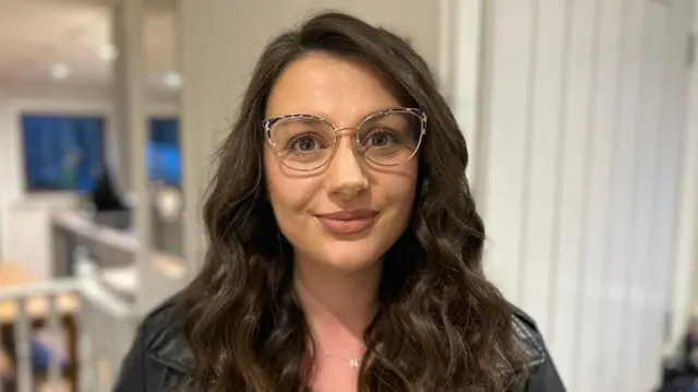 Sarah Goddard tem cabelos castanho-escuros ondulados e sorri para a câmera; ela usa óculos e jaqueta preta de couro