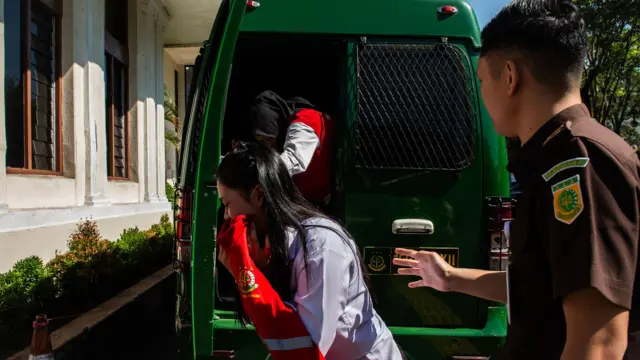Dua orang perempuan dengan kemeja putih turun dari mobil tahanan berwarna hijau tua, seorang petugas pengadilan berdiri di belakang mobil.