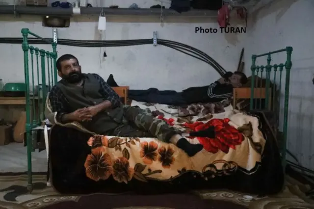 Предположительно подвальное помещение, с кроватями на которых лежат люди.