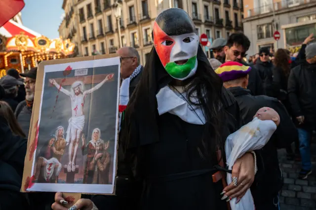 سيدة ترتدي قناعاً بلون علم فلسطين، وتحمل دمية لطفل في كفن على يدها اليسرى، وفي يديها اليمنى لوحة للمسيح مصلوباً وهو يرتدي العقال الفلسطيني وحوله مجموعة من الفلسطينيين المكلومين