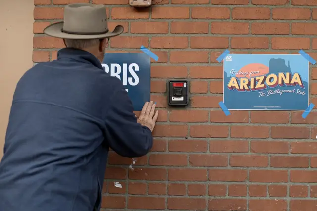 Uma pessoa de costas cola uma placa de campanha ao lado de outra escrito Arizona.
