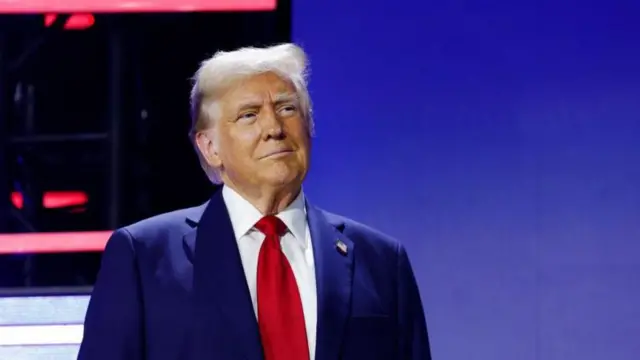 Donald Trumpo, um homem branco idoso de terno com bronzeado artificial e cabelos grisalhos
