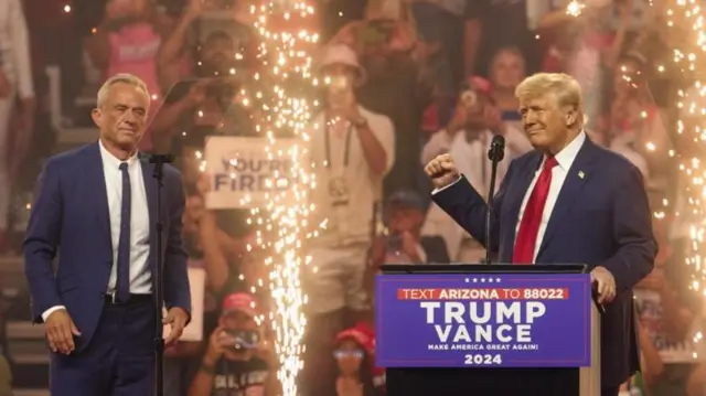 Robert F Kennedy Jr. e Trump perto de palanque, sorrindo, com fogos e apoiadores atrás