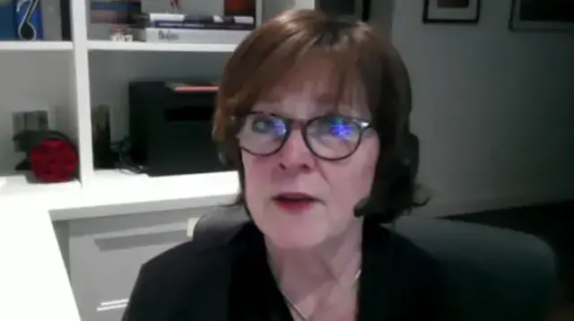 Mulher branca de óculos com estante ao fundo