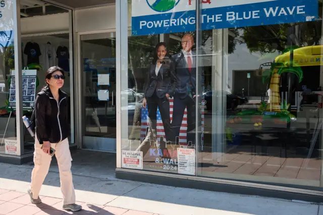 Mulher passando em frente a escritório democrata em Los Angeles; dentro, há foto de Harris e Walz em tamanho real