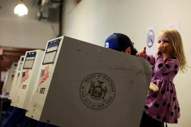 Uma criança aparece no colo com um adulto em uma cabine de votação