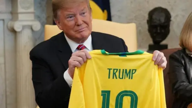 Trump, um homem branco idoso de cabelo claro e bronzeado artificial, segura uma camiseta verde e amarela com o número 10 nas costas e a palavra Trump