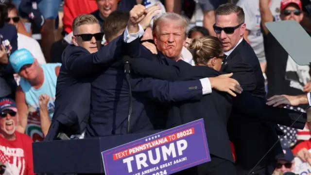 Trump sangrando no rosto e levantando o punho, rodeado por seguranças