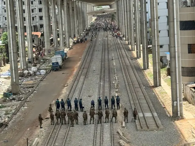 ঢাকার মহাখালী থেকে তেজগাঁও যাবার রেললাইনের ওপর জড়ো হয়েছে একদল মানুষ। তাদের মুখোমুখি কিছুটা দূরে মহাখালী মোড়ের কাছে অবস্থান নিয়ে রয়েছে পুলিশ ও সেনাবাহিনীর সদস্যরা। দুপুর ১২টার ছবি