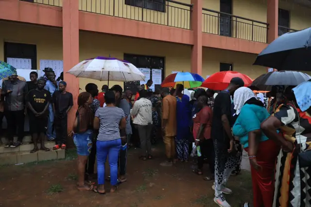 Pipo wey dey wait to cast dia vote for di Edo state govnorship election hold umbrella as rain dey fall
