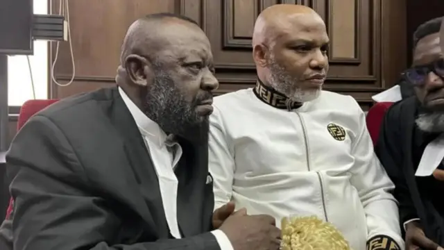 Ejimakor yi uwe ojii ndị ọkaiwu nọrọ Kanu yi uwe ọcha n'akụkụ n'ime ụlọikpe