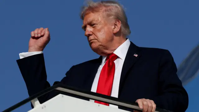 Дональд Трамп в красном галстуке поднимает кулак вверх, стоя на трапе самолета