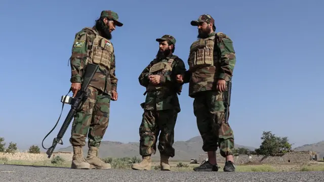 د طالبانو درې سرتېري په پوځي درېشیو کې چې وسلې ورسره او چانټې یې اغوستې دي، پر سړک ولاړ دي او شا ته یې کورونه برېښي.