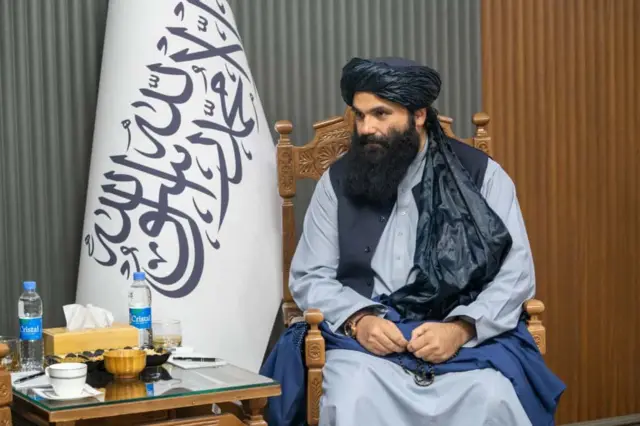 سراج الدین حقاني توره بګړۍ وهلې، شینبخون څادر یې په غېږ کې اچولی، تسبې اړوي او اوبرنګه کالیو باندې یې تور واسکټ اغوستی. څنګ ته یې د طالبانو بیرغ ولاړ دی.